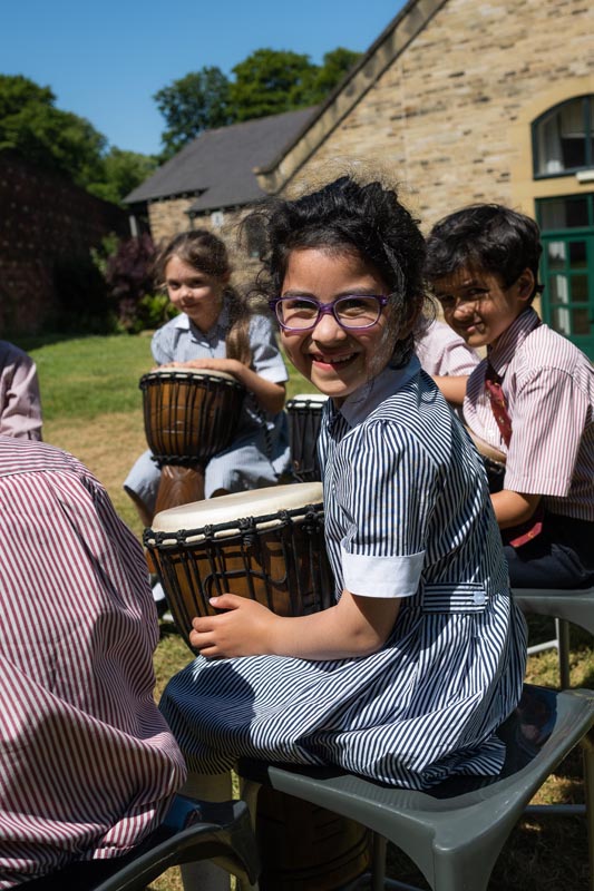 Image of pupils in a circle with drums on their knees. Girl at the front is smiling to the camera