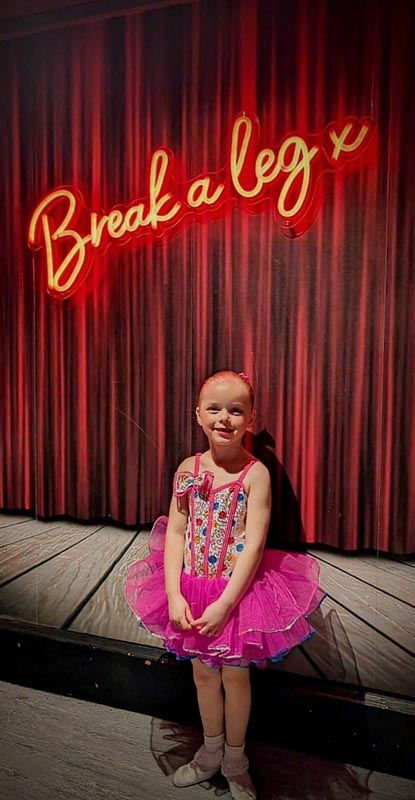 Young ballerina in pink tutu poses in front of Break a Leg sign