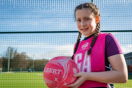 A girl in a pink GA vest holding a pink netball smiling to the camera