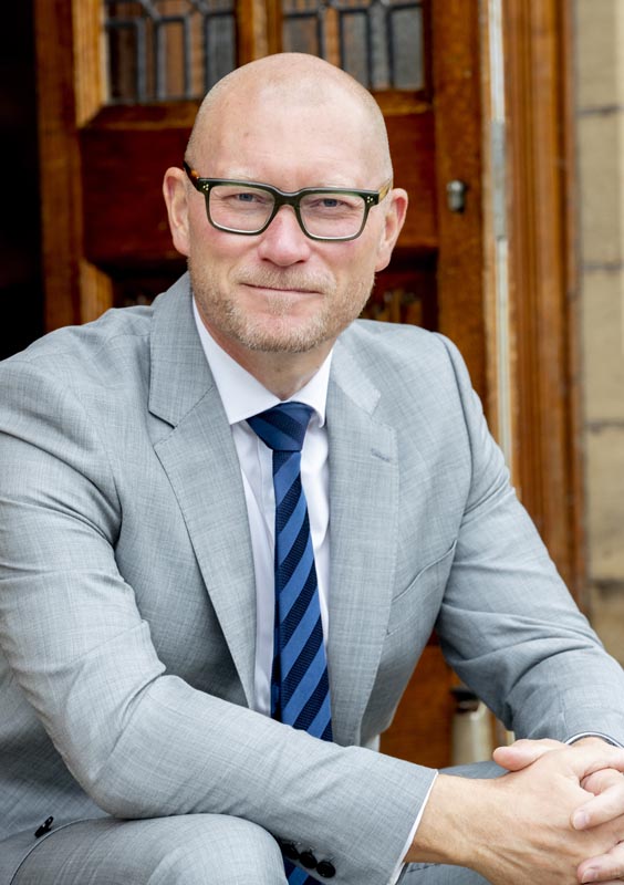 Dr Simon Hinchliffe - BGS Headmaster, wearing a grey suit and a blue striped tie, seated in front of a wooden door, smiling at the camera.