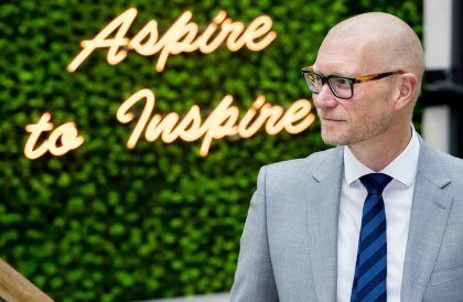 Headmaster Dr Simon Hinchliffe in a grey suit with a blue tie, standing in front of a neon sign that reads 'Aspire to Inspire' on a green hedge.