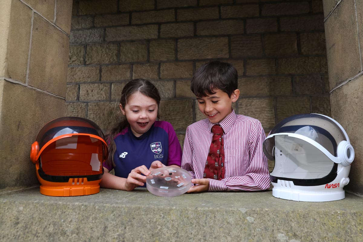 Two children leaning on a stone archway holding pieces of the moon alongside two astronaut helmets.